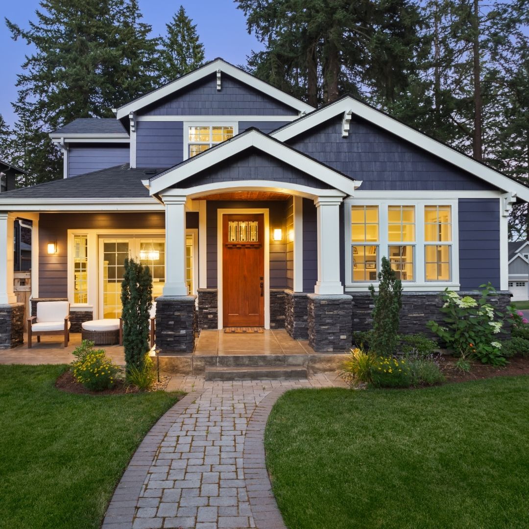A modern two-story home with a blue and wood facade, a lit interior, and well-maintained landscaping including a grass lawn and white stone pavers at dusk.