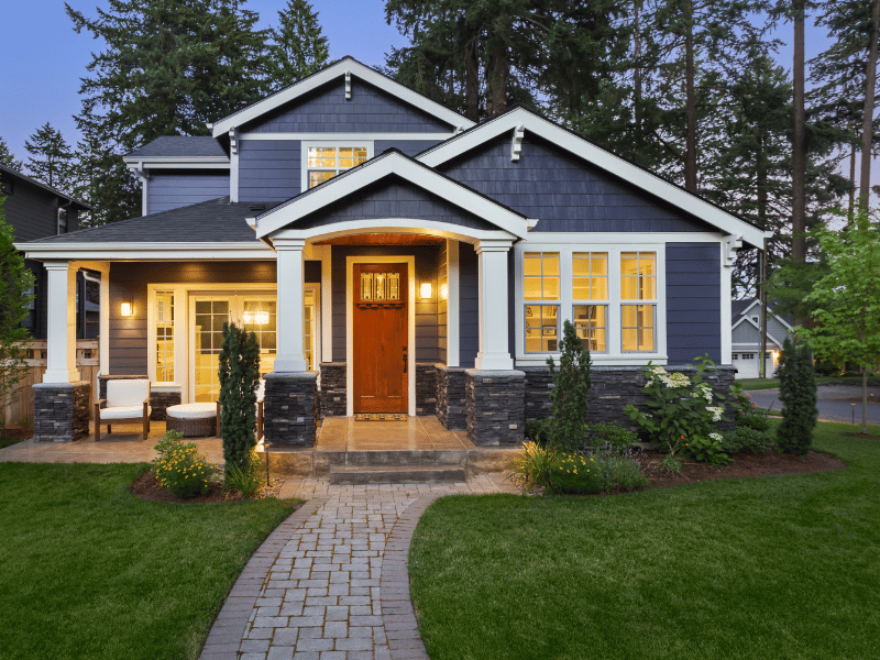 A modern two-story home with a blue and wood facade, a lit interior, and well-maintained landscaping including a grass lawn and white stone pavers at dusk.