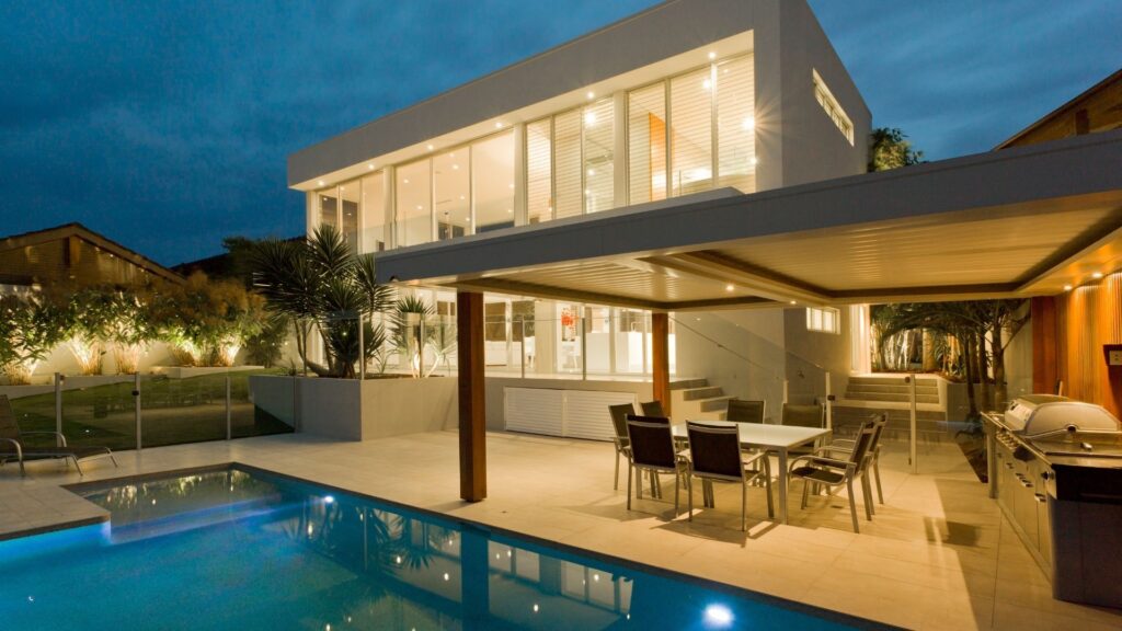 Modern luxury backyard hardscaping in Boston featuring a large stone tile patio, a custom outdoor kitchen with a professional grill, and a sleek swimming pool.