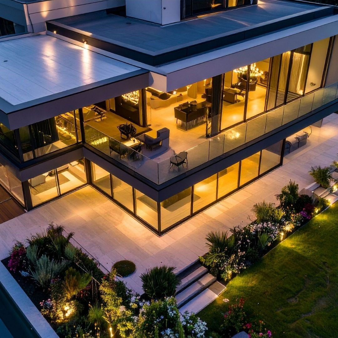 Elevated evening view of a luxury modern villa with floor-to-ceiling glass walls, warm interior lighting, a spacious balcony, and manicured garden landscaping.